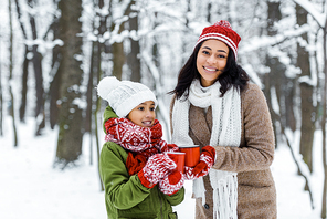 attractive african american mother and cute daughter holding red cups of tea and staying near trees in snowy forest