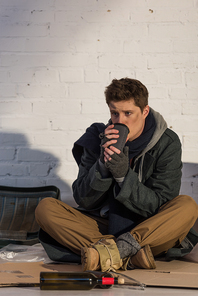 sad homeless man drinking from paper cup while sitting on cardboard by brick wall