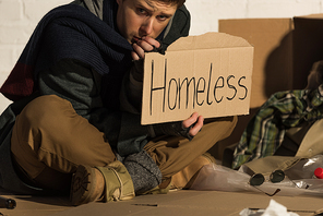 depressed man sitting on cardboard and holding card with homeless handwritten text