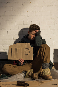 despaired homeless man sitting by brick wall and holding piece of cardboard with help inscription