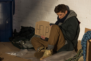 depressed homeless man holding piece of cardboard with help inscription while sitting on trash dump