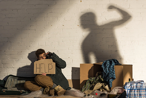 frightened homeless man holding card with help lettering while sitting by brick wall with shadow of man raising hand with fist