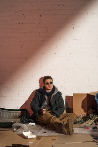 homeless man surrounded by rubbish sitting by white brick wall