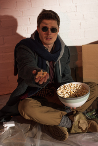 homeless man holding popcorn in raised hand while sitting by white brick wall