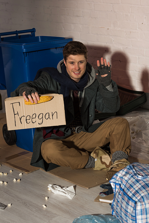 smiling homeless man waving hand while holding cardboard card with freegan inscription and corn cob