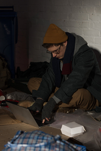 homeless man using laptop while sitting by white brick wall surrounded by rubbish