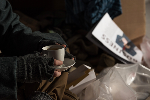 selective focus of homeless man holding coffee cup while sitting on rubbish dump