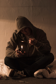 junkie man sitting in dark room and heating spoon with heroin on lighter