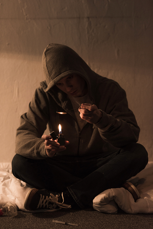 junkie man sitting in dark room and heating spoon with heroin on lighter