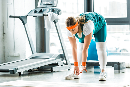 concentrated overweight woman stretching near fitness mat and treadmill in gym