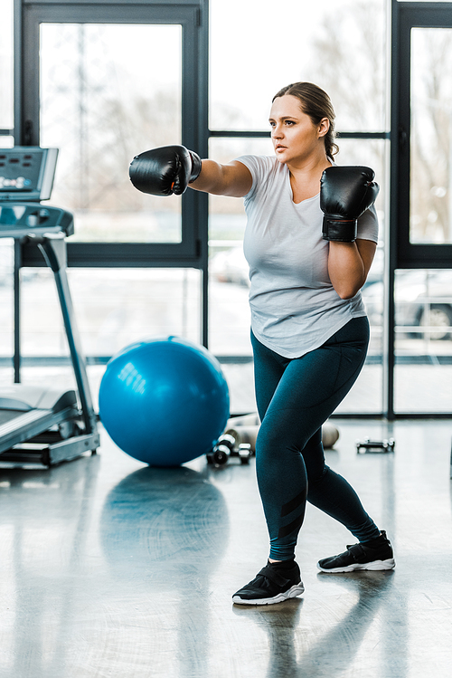 serious plus size girl wearing boxing gloves and practicing kickboxing in gym