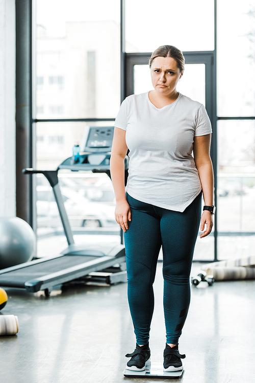 upset overweight woman standing on scales in gym