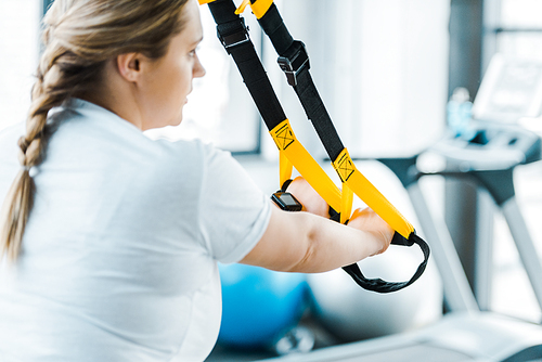 selective focus of overweight woman training arms with suspension straps in gym