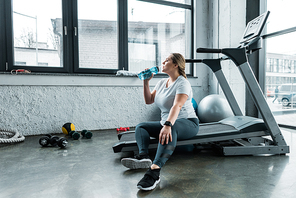 tired plus size woman sitting on treadmill and drinking water from bottle