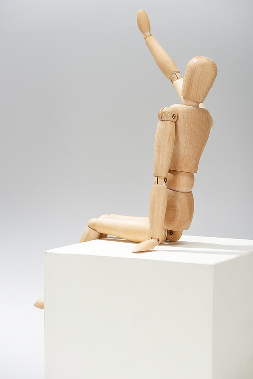 Wooden puppet with outstretched hand on white cube on grey background