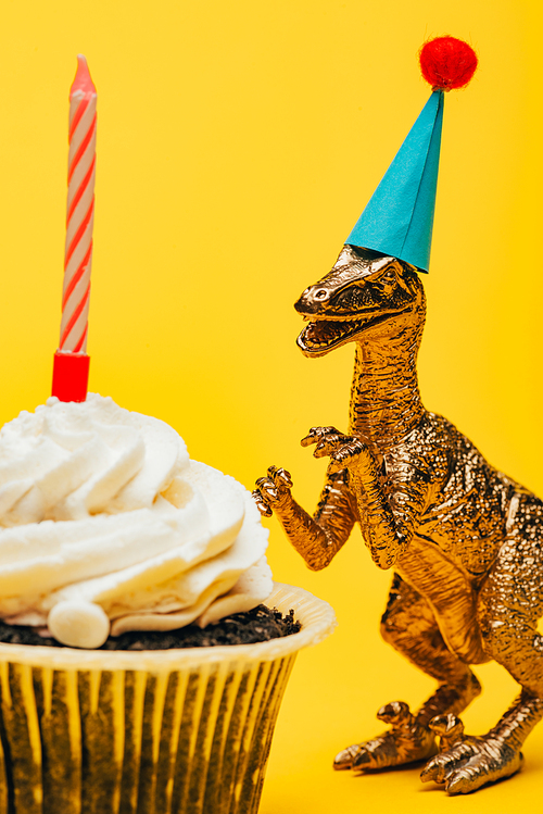 Selective focus of toy dinosaur in party cap beside cupcake with candle on yellow background