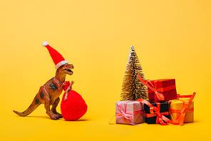 Toy dinosaur in santa hat and sack beside pine with gifts on yellow background