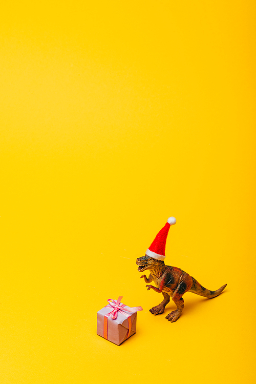 Toy dinosaur in santa hat with gift on yellow background