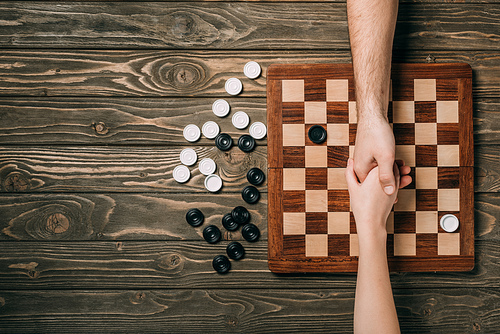 Cropped view of man and woman shaking hands above checkerboard with checkers on wooden background