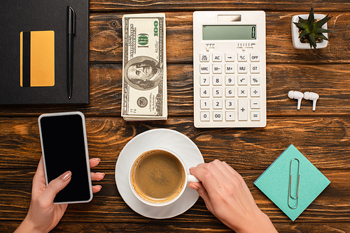 cropped view of businesswoman holding smartphone and coffee cup near dollar banknotes, calculator, wireless earphones and stationery on wooden desk