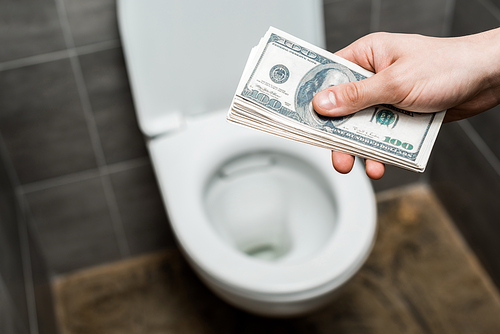 cropped view of man holding dollar banknotes near toilet bowl in modern restroom with grey tile