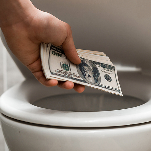 cropped view of man holding dollar banknotes near toilet bowl in modern restroom with grey tile