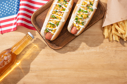 top view of delicious hot dogs with corn, green onion and mayonnaise near french fries, american flag and beer on wooden table