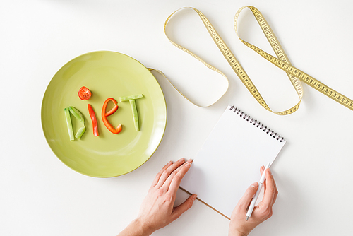 Top view of woman writing in notebook near measuring tape and plate with diet lettering from vegetable slices on white background