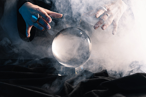 Cropped view of female hands above crystal ball with smoke on dark