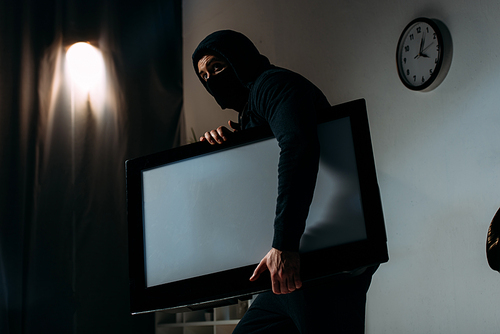 Robber in black mask stealing flat-screen tv with blank screen from apartment