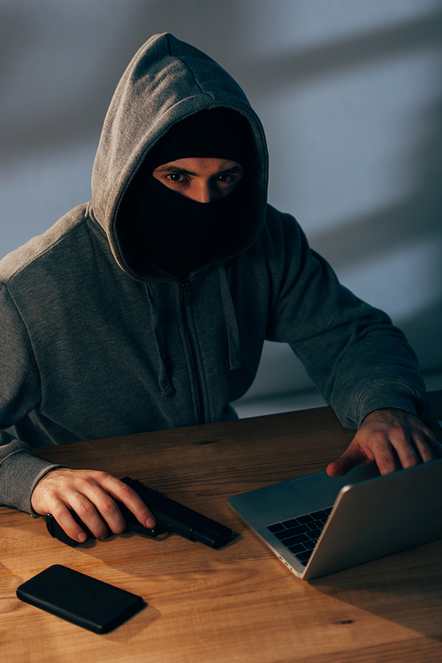 Criminal in mask holding gun and using laptop in dark room