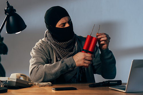 Terrorist in mask holding dynamite while sitting at table in dark room