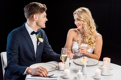 happy young couple sitting at served table with glasses of champagne and looking at each other isolated on black