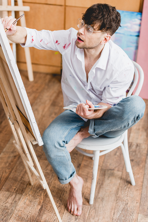 selective focus of handsome artist painting on canvas while sitting on chair in painting studio