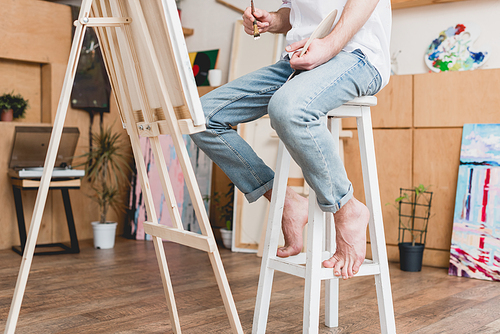 partial view of barefoot artist sitting on high chair in painting studio