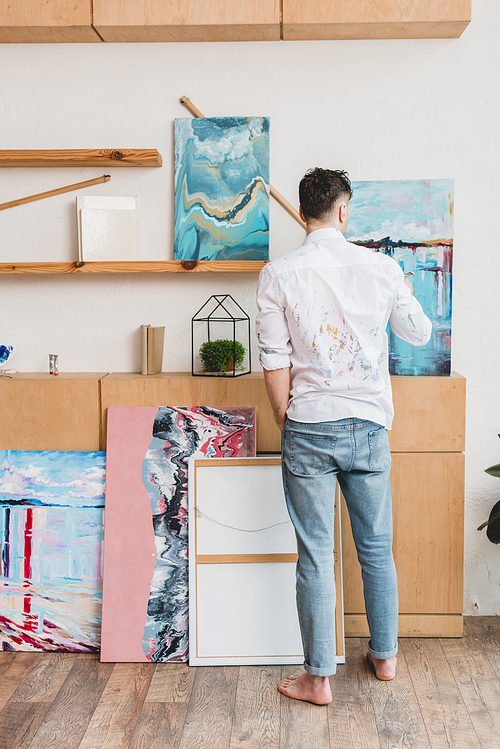 back view pf barefoot artist in white shirt and blue jeans standing in painting studio