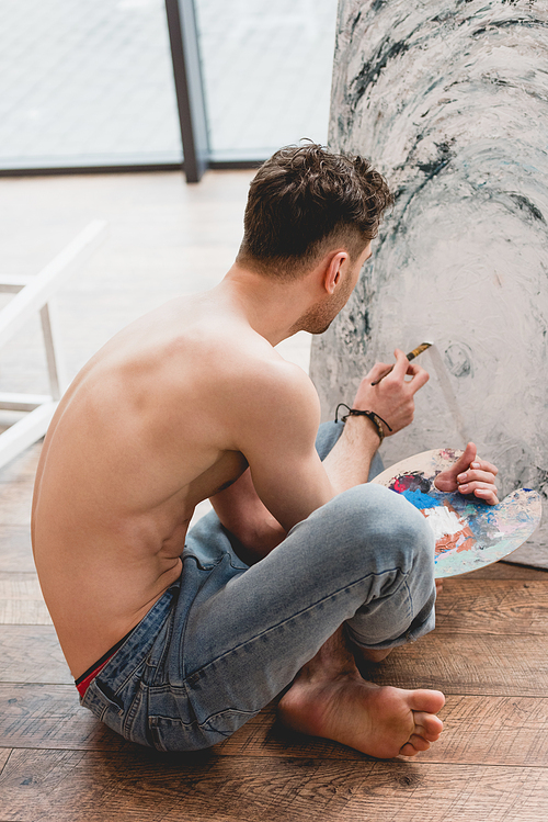 half-naked artist sitting with crossed legs and painting picture in gallery