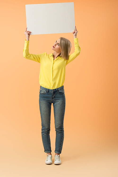 Full length view of irritated blonde girl in jeans holding blank placard on orange background