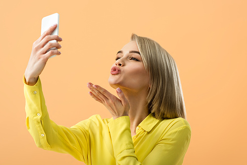 Lovely blonde girl sending air kiss while taking selfie isolated on orange