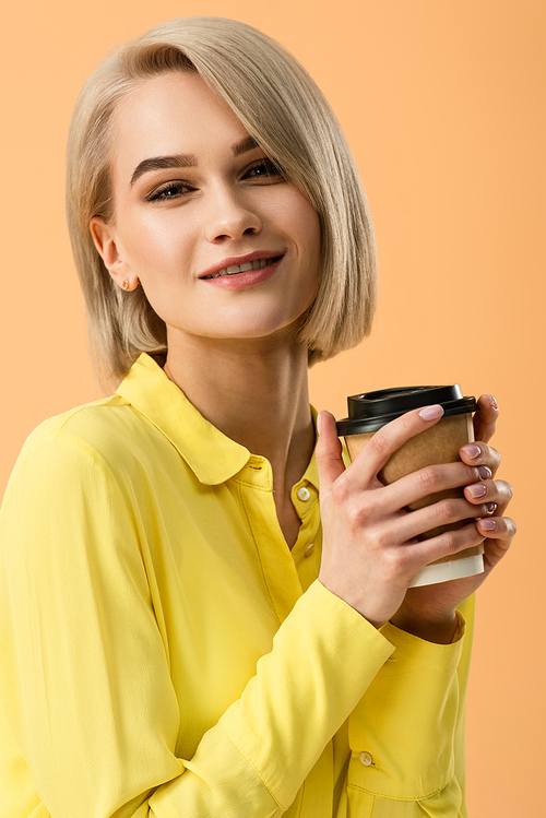 Smiling beautiful blonde girl holding paper cup of coffee isolated on orange