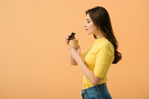 Side view of relaxed brunette woman holding paper cup of coffee isolated on orange