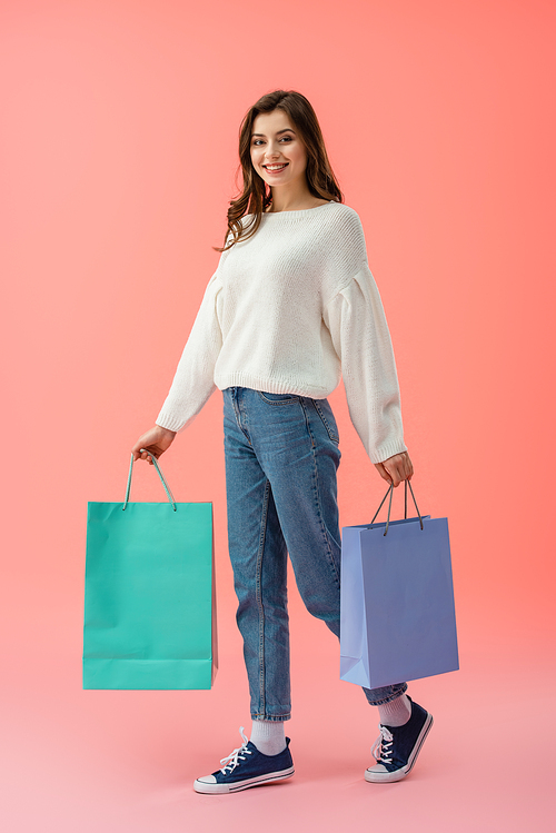 full length view of woman in white sweater and jeans holding shopping bags on pink background