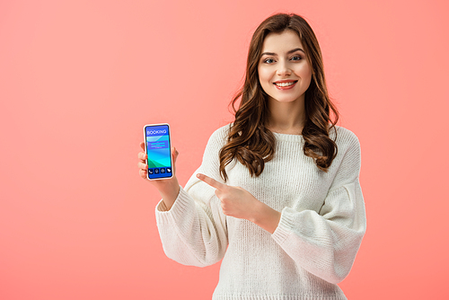 woman in white sweater pointing with finger at smartphone with booking app on screen isolated on pink