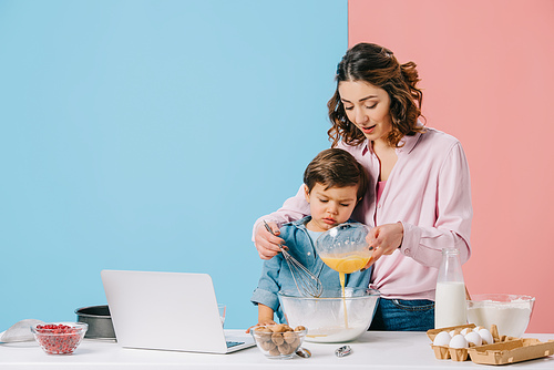 mother adding whipped eggs to bowl while cooking together with little son on bicolor background