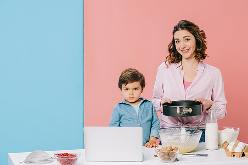 mother holding baking form while cooking together with little son at kitchen table with products, cooking utensils and laptop on bicolor background