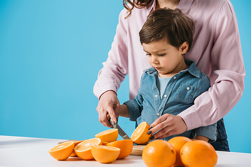 cute little boy cutting oranges together with mother isolated on blue