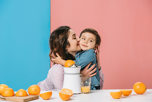 happy mother hugging adorable smiling little son by kitchen table with fresh oranges and juicer on bicolor background