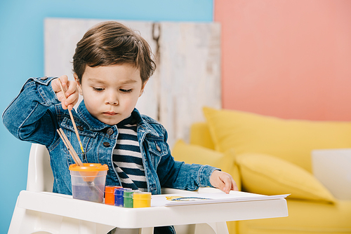 cute little boy choosing painting brush while sitting on highchair with watercolor paints on table