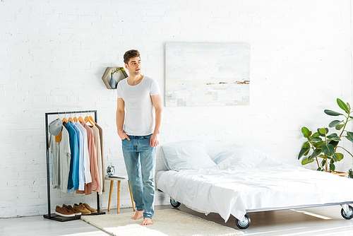 handsome man in white t-shirt and jeans standing near bed and clothes rack in bedroom