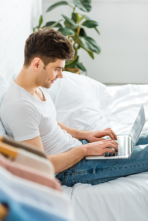 selective focus of handsome man in white t-shirt and jeans sitting on bed and typing on laptop in bedroom